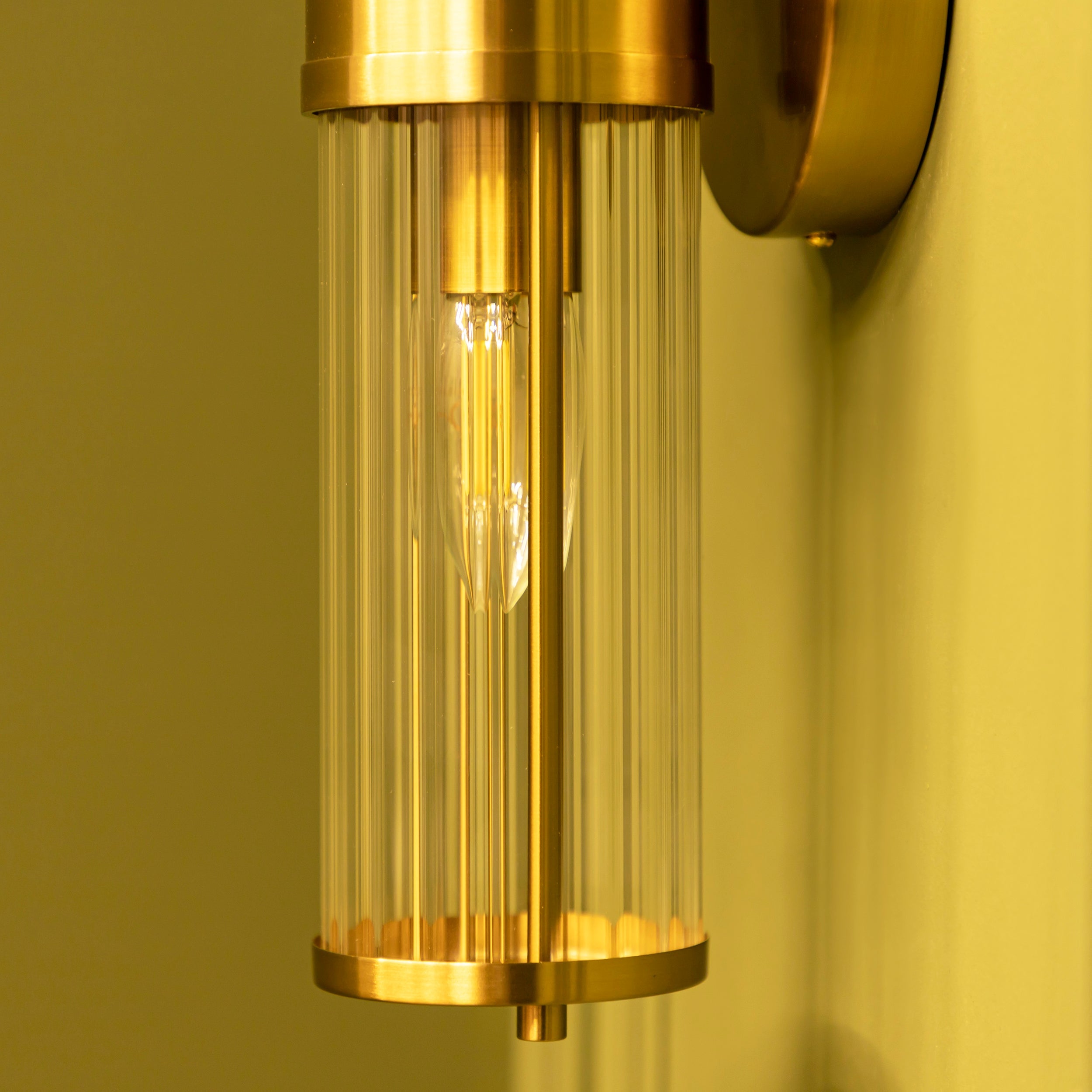 Gold wall-mounted light fixture with clear glass cover on a yellow background