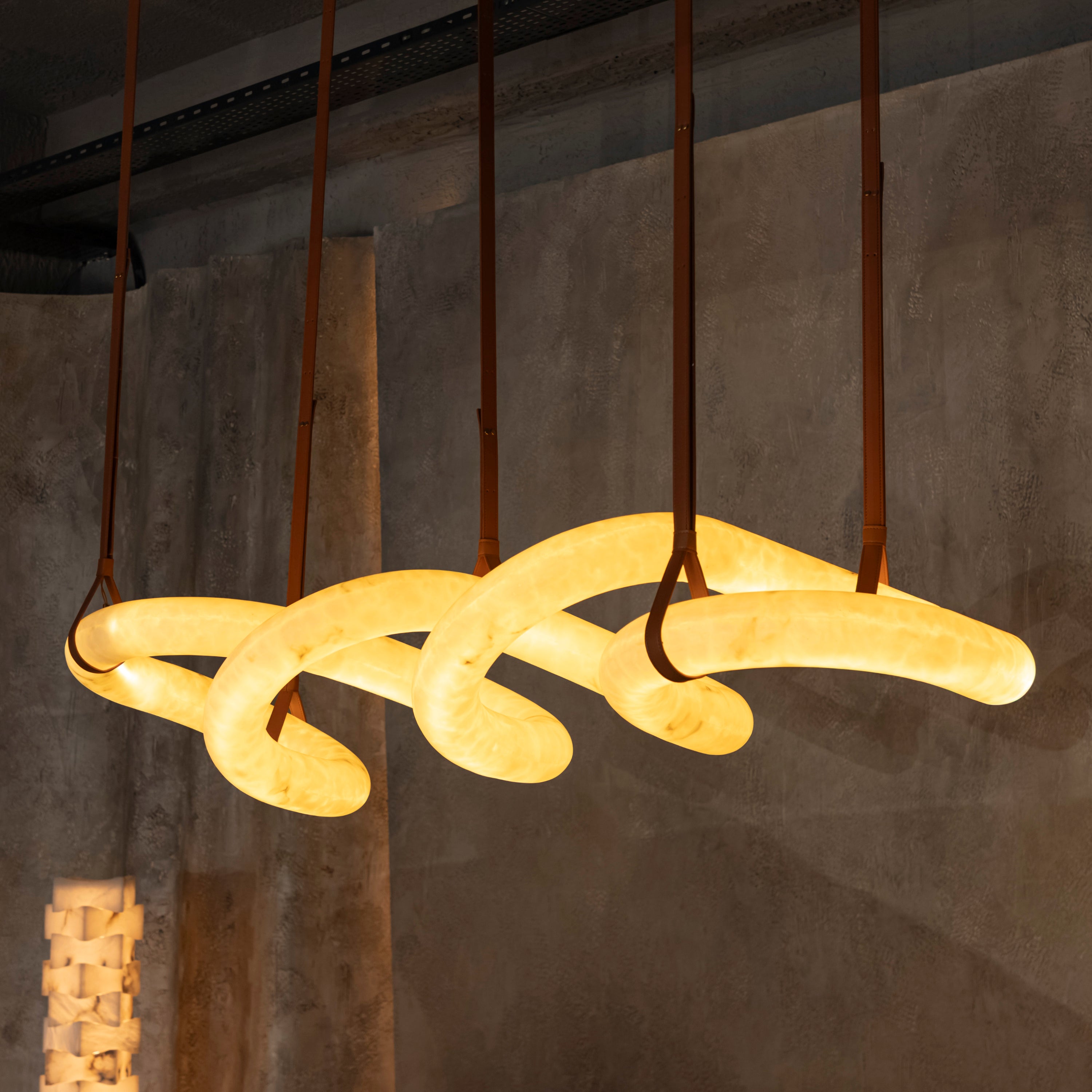 A modern multi-arm pendant light with a glowing warm yellow organic curve design, suspended by brown cords against a textured concrete wall.