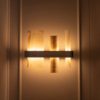 A modern wall-mounted light fixture with three vertical translucent amber panels and a horizontal dark metal bar, shown lit against a warm beige paneled wall.