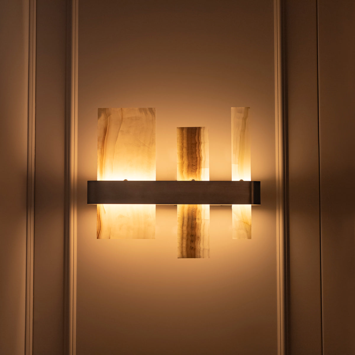 A modern wall-mounted light fixture with three vertical translucent amber panels and a horizontal dark metal bar, shown lit against a warm beige paneled wall.