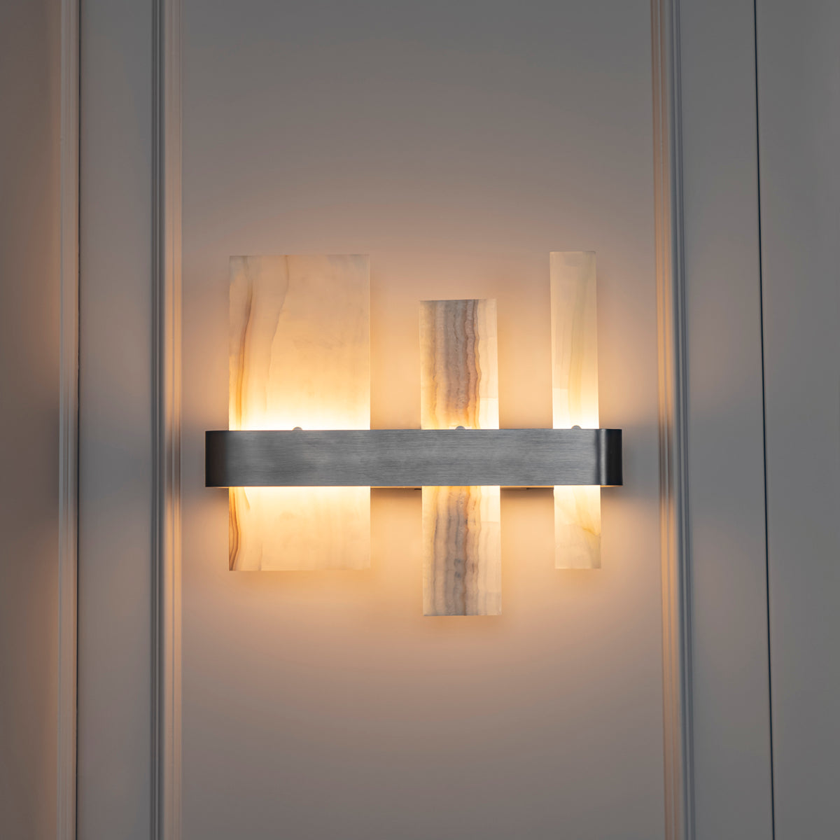 A modern wall sconce light fixture with three vertical translucent stone or acrylic panels and a horizontal brushed metal bar, mounted on a light gray paneled wall.