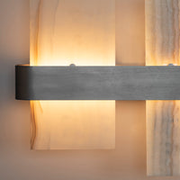 A modern wall sconce featuring a brushed metal horizontal bar mounted on vertical textured stone panels that glow with warm light.