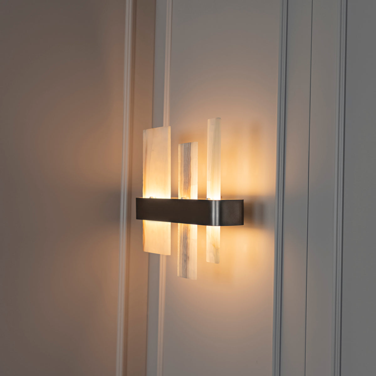 A modern wall-mounted sconce with a brushed metal rectangular base and three translucent rectangular glass shades, glowing warm light against a paneled wall.