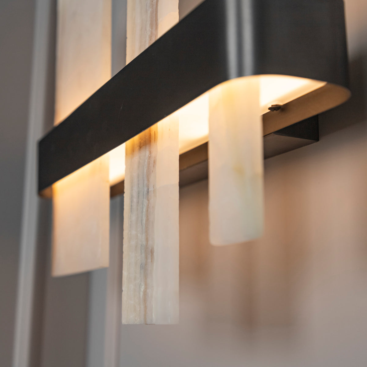Modern wall-mounted light fixture with a matte black metal frame and vertical translucent stone or acrylic panels, shown lit against a softly blurred interior background.
