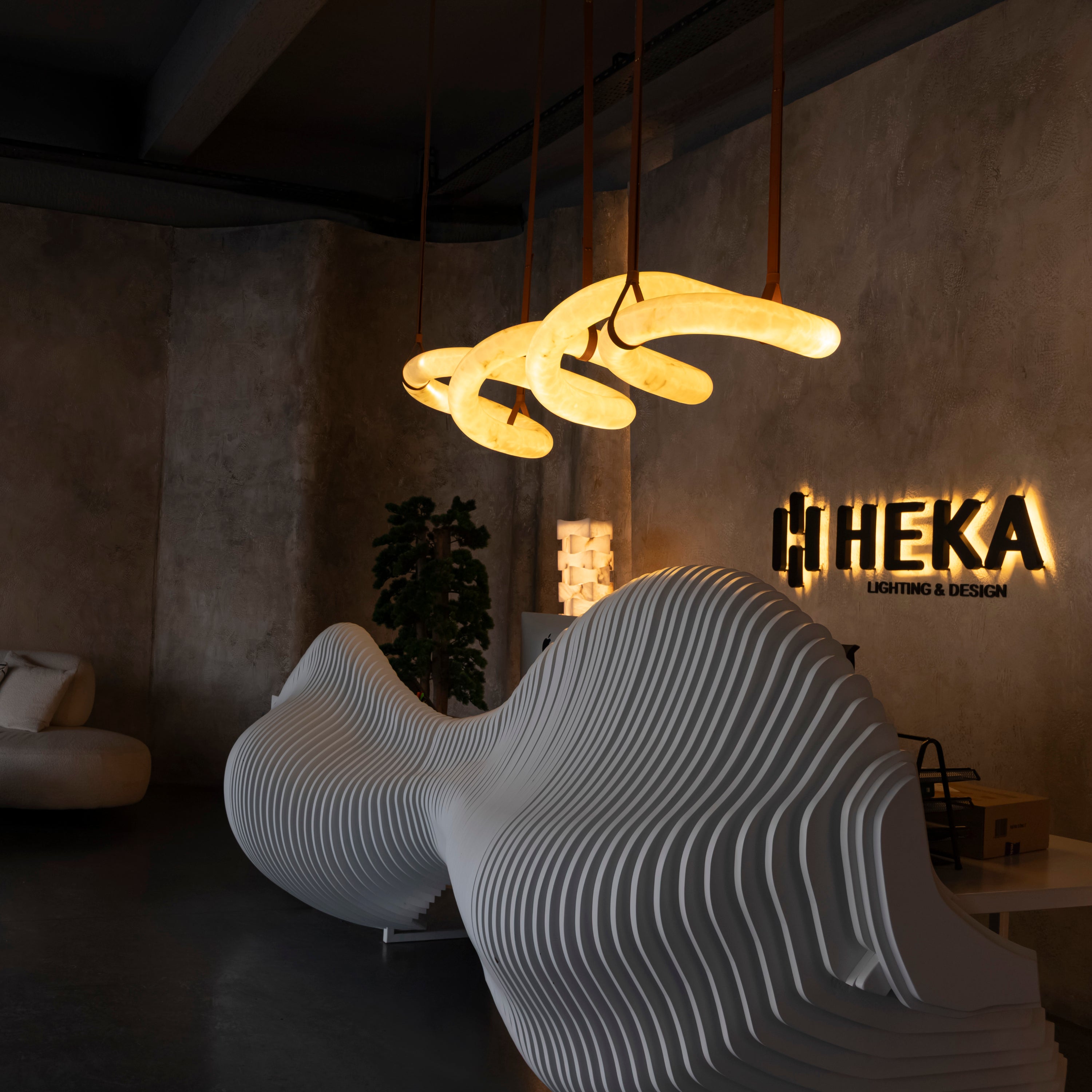 A modern wavy pendant light with a glowing warm yellow-white curved tube design hangs in a dimly lit industrial-style room. A large sculptural ribbed white sofa sits in the foreground, with a textured concrete wall behind showing a black illuminated 'HEKA Lighting & Design' sign.